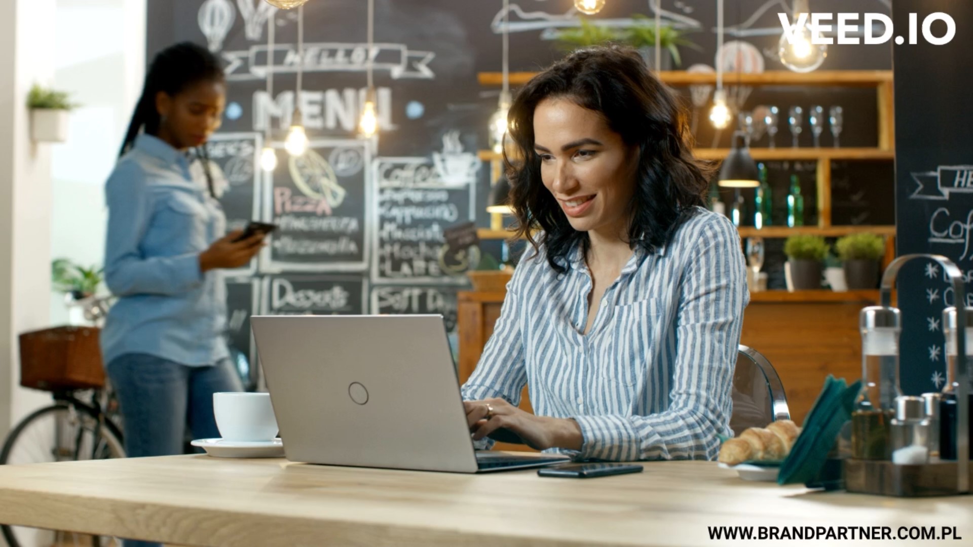 Woman working on laptop another woman in background using phone